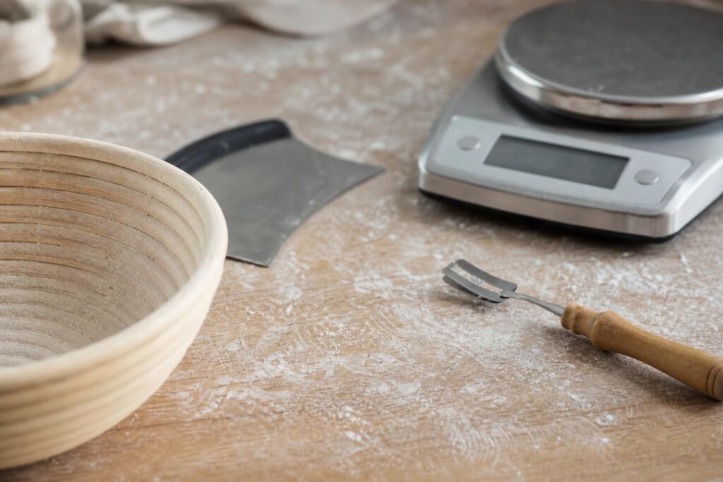 Matériel pour faire son pain maison : balance, corne, banneton en rotin et lame posés sur un plan de travail fariné